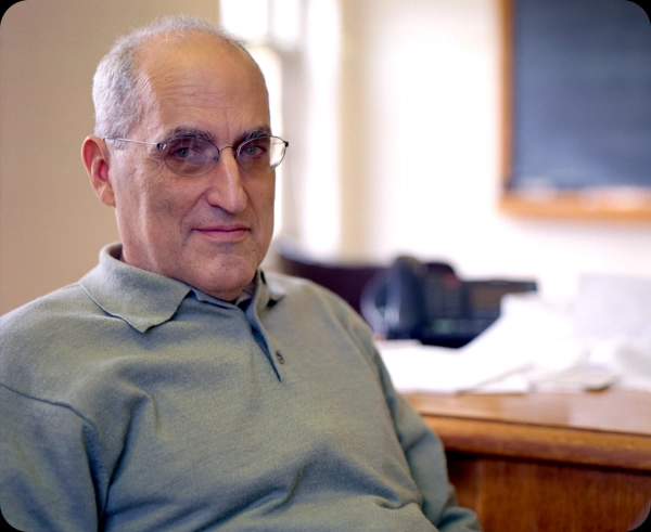 В офисе сидит мужчина в очках и зеленой рубашке.