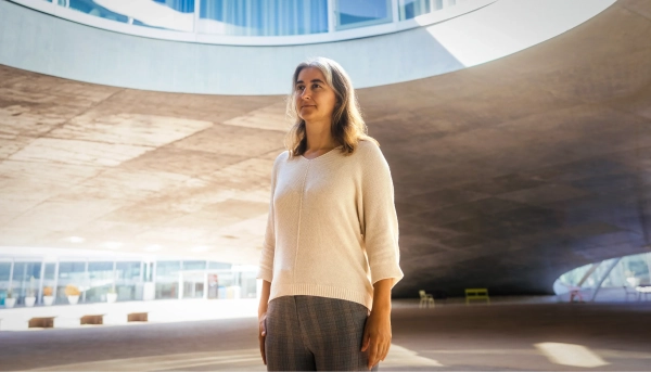 Женщина в белом свитере стоит перед крупным архитектурным сооружением.