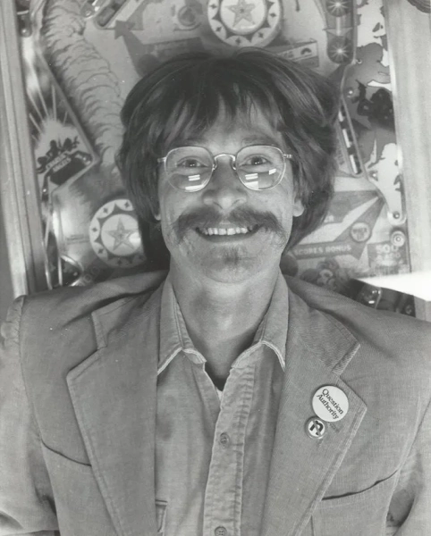 Грибной гроб стал первым примером «зеленых похорон» в США. 3 Чёрно-белое фото мужчины в очках, улыбающегося в камеру. У него усы, а на куртке — значок с надписью «Подвергайте сомнению авторитет».