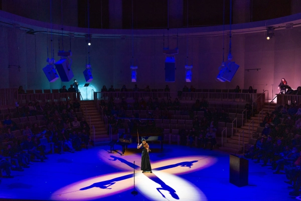 Женщина держит смычок высоко поднятым, играя на скрипке в центре сходящихся лучей прожекторов таким образом, что от нее отходят четыре тени под углом 90 градусов.