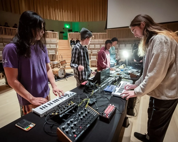 Студенты на сцене стоят за длинным столом с клавиатурами и другими контроллерами.