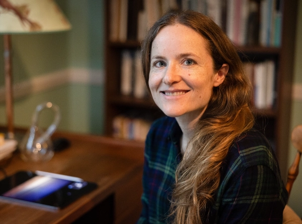 Молодая женщина с каштановыми волосами читает книгу за столом.