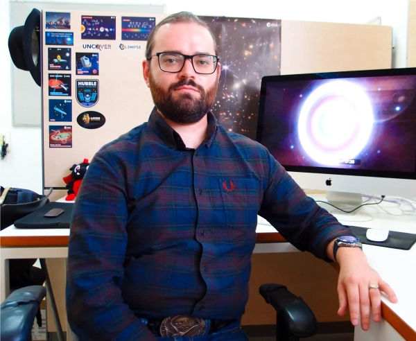 Одна «голая» чёрная дыра переписывает историю Вселенной 4 Перед столом сидит бородатый мужчина в темно-синей рубашке.