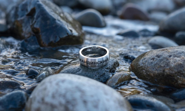 Prompt: Стильное рекламное фото. Крупный план. Среди мокрых мрачных скал и камней в медленном ручье ледяной воды и в луче света лежит серебряное мужское кольцо с надписью КАНДИНСКИЙ
