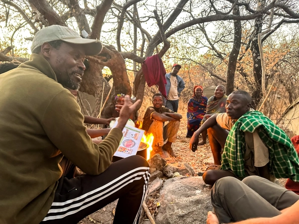 Дуглас беседует у костра с хаздой, держа в руках инфографику.