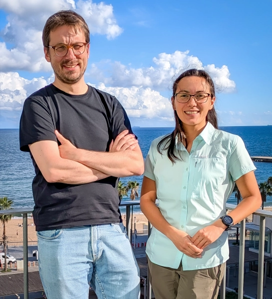 Мужчина и женщина стоят вместе на балконе, на фоне моря. Мужчина слева улыбается. У него короткие каштановые волосы, темная футболка и джинсы. Он в очках, улыбается, скрестив руки на груди. Женщина справа ниже ростом, тоже в очках, улыбается. На ней светло-голубая рубашка с короткими рукавами и светло-коричневые брюки.