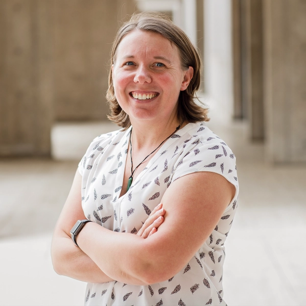 Женщина смотрит в камеру, скрестив руки на груди, и улыбается. Её каштановые волосы до плеч, лёгкое узорчатое платье, на левом запястье – ожерелье и часы.