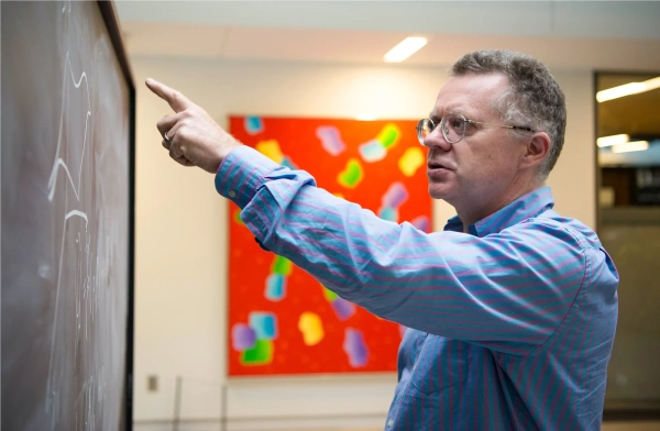 Мужчина в синей рубашке с красными полосками указывает на доску.