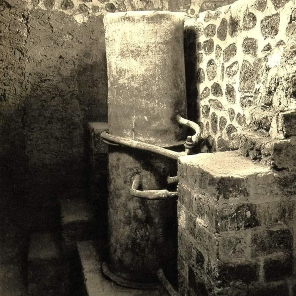 "Необыкновенная находка, найденная при раскопках виллы Пизанелла в Боскореале, недалеко от Помпей. К сожалению, это поселение не было объектом систематических раскопок, а только частичных исследований.Эти быстрые раскопки, проведенные без какой-либо научной систематики, имели целью извлечение драгоценных находок, которые сегодня разбросаны в различных музеях по всему миру.Первые свидетельства о вилле делла Пизанелла датируются ноябрем 1868 года, когда Модестино Пульцелла, прослеживая фундамент стены, обнаружил некие конструкции. Продолжая раскопки, была найдена мозаика. Затем раскопки пришлось прекратить, поскольку владелец соседней земли Винченцо Де Приско указал на опасность ущерба для его земли. Только в сентябре 1894 года раскопки были возобновлены, которые продолжались до июня 1895 года. На этот раз именно по инициативе Де Приско, который, очевидно, больше не боялся «ущерба».Так всплыло на свет знаменитое «сокровище Боскореале», сенсационно проданное за границу из-за пробела в законодательстве о защите культурного наследия. Был еще один перерыв, который длился около года, прежде чем раскопки возобновились в мае 1896 года.Так на свет появились римские бани и котел, найденные нетронутыми, со всеми их трубами. Затем Де Приско провел реставрацию части виллы, превратив ее в своего рода музей.И именно во время существования «Музея Де Приско» братья Алинари получили возможность сделать эту необычную фотографию"