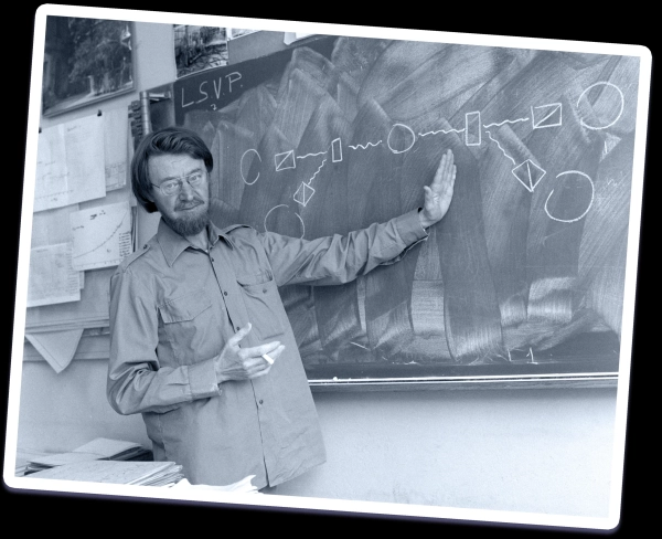 Черно-белая фотография мужчины с усами и бородой, показывающего на рисунок на доске, на котором изображен план эксперимента.