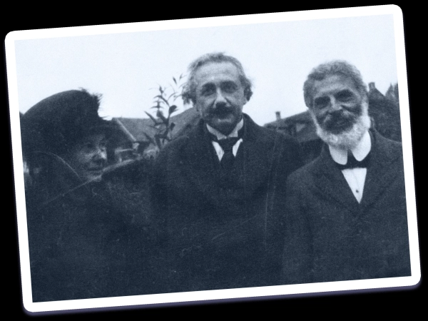Черно-белая фотография трёх человек, стоящих перед домом и дружелюбно улыбающихся. Мужчина в центре — Эйнштейн.