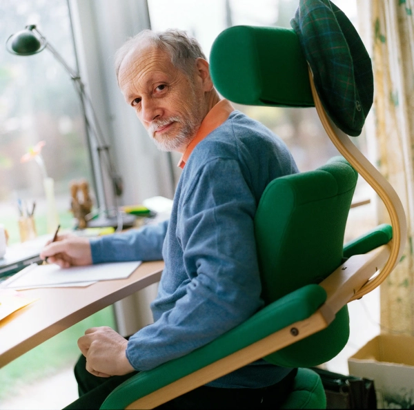 Мужчина в синем свитере сидит в зеленом кресле за столом в залитой солнцем комнате.