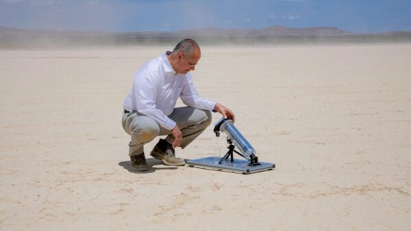 Человек в пустыне смотрит в телескоп на треноге, солнечный день, ясное небо.