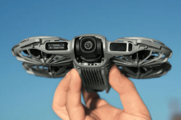 Рука держит компактный дрон с камерой на фоне голубого неба.