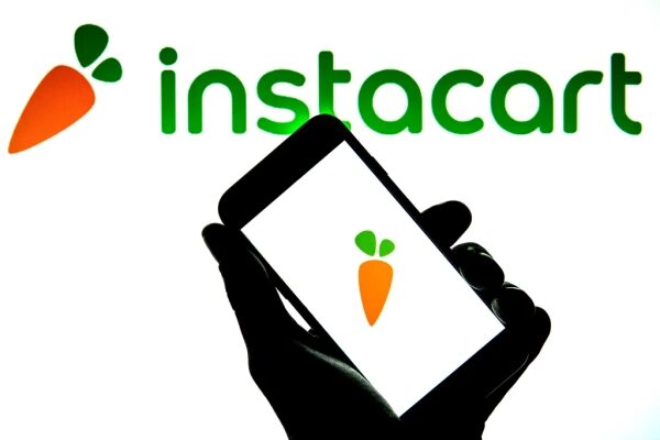Логотип Instacart на экране смартфона, поднятого на фоне логотипа на стене.