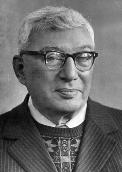 Пожилой человек в костюме и очках на черно-белом портрете.