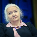 Пожилая женщина с седыми волосами и серьезным выражением лица на фоне синего цвета.