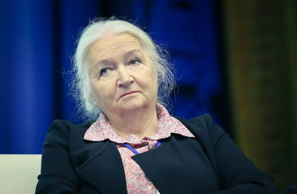 Пожилая женщина с седыми волосами и серьезным выражением лица на фоне синего цвета.