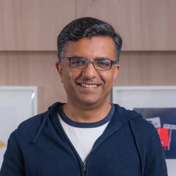 Мужчина в очках и толстовке синий на фоне деревянной стены в офисе улыбается.