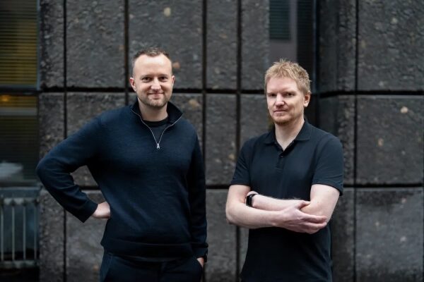 Два мужчины в чёрной одежде стоят у серой стены на улице.