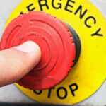 Палец нажимает красную кнопку аварийной остановки на пульте управления.