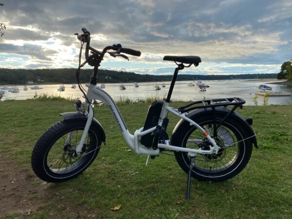 Электровелосипед на берегу реки с лодками и облачным небом на фоне.