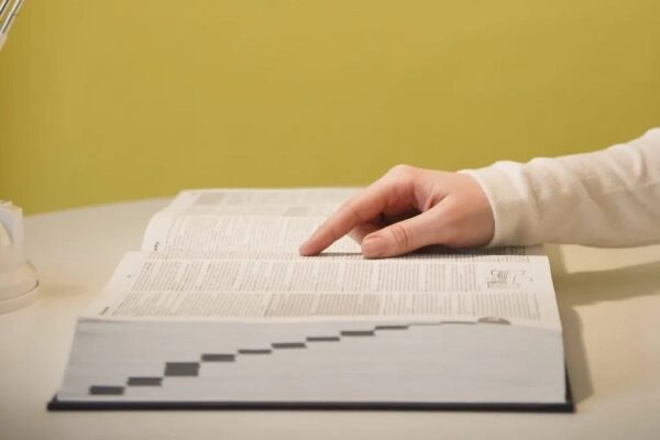 Рука листает толстую книгу на белом столе перед жёлтой стеной.