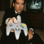 Человек в смокинге держит контроллер Nintendo 64, на экране игра.