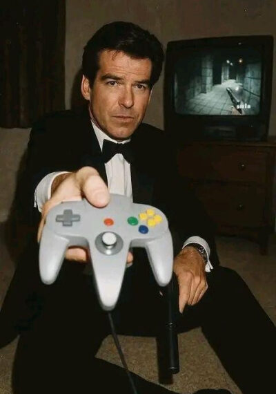 Человек в смокинге держит контроллер Nintendo 64, на экране игра.