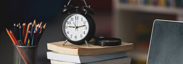 Чёрный будильник на книгах, карандаши в стакане, ноутбук на рабочем столе.