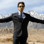 Человек в костюме стоит в пустыне на фоне гор с разведенными руками.