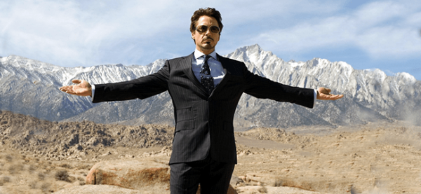 Человек в костюме стоит в пустыне на фоне гор с разведенными руками.