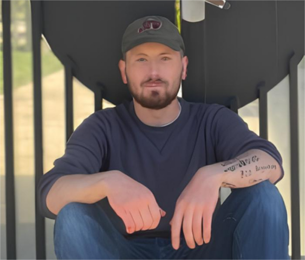 Мужчина в кепке и джинсах сидит на улице, на руке татуировка.