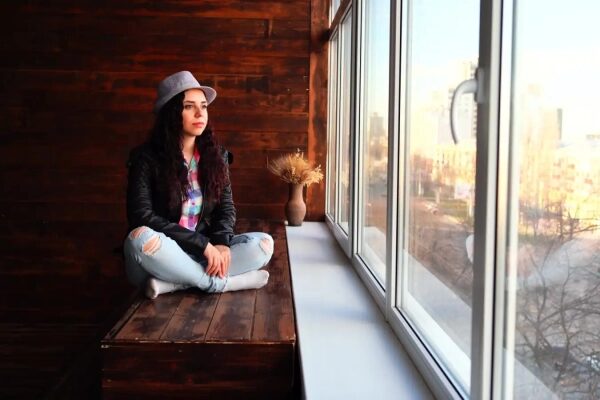 Девушка в шляпе сидит у окна на деревянной лавке, задумчиво смотрит вдаль.