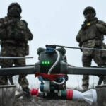 Дрон с грузом на фоне военных в камуфляже на открытой местности.