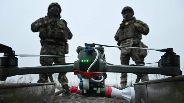 Дрон с грузом на фоне военных в камуфляже на открытой местности.