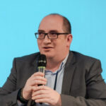 Мужчина в костюме говорит в микрофон на фоне голубой стены.