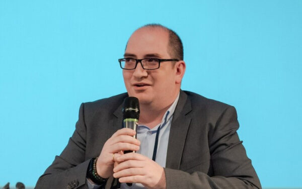 Мужчина в костюме говорит в микрофон на фоне голубой стены.