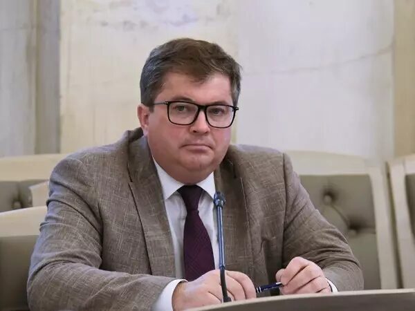 Мужчина в очках и костюме за столом с микрофоном на конференции.