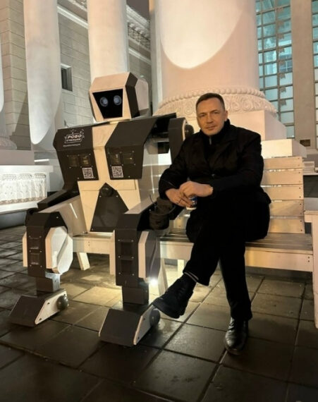 Человек сидит рядом с роботом на скамейке у колонн ночью.