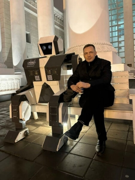 Человек сидит рядом с роботом на скамейке у колонн ночью.