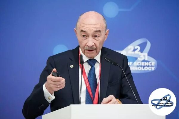 Мужчина выступает на конференции науки и технологий, держит указку у синего фона.