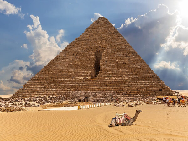 Пирамида в Египте, верблюд в пустыне на фоне голубого неба и облаков.