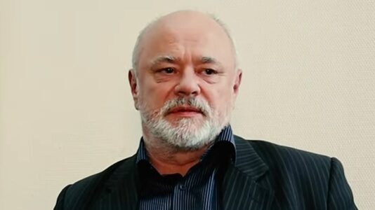 Мужчина с бородой в костюме на фоне светлой стены смотрит вперед.