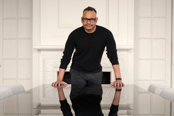 Человек в черной одежде стоит у стола в светлой комнате.