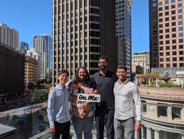 Четыре мужчины стоят на улице в деловом районе города, держа табличку "On Me".