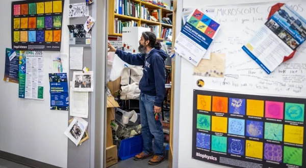 Ману Пракаш стоит в своем кабинете; по обе стороны двери висят плакаты, посвященные «экономной науке» и «биофизике».