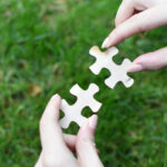 Руки держат части пазла на фоне зеленой травы.