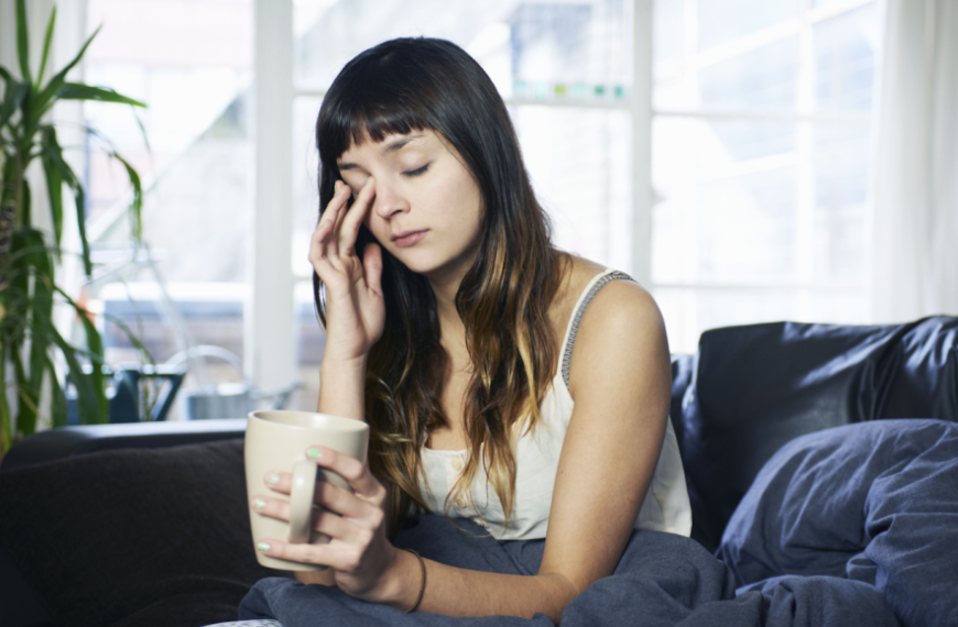 Девушка с кружкой в руках сидит на диване, просыпается и потирает глаза.