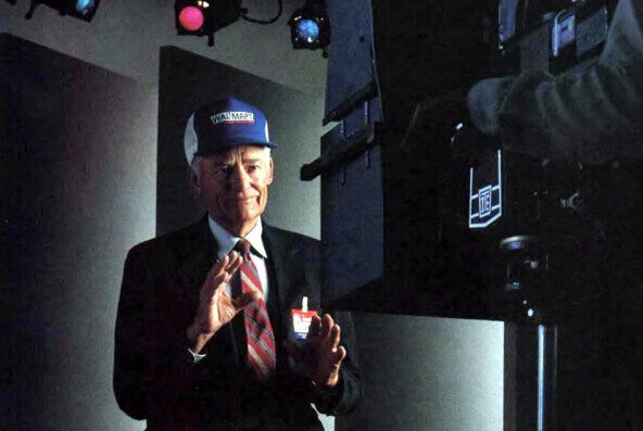 Человек в костюме и бейсболке позирует перед камерой в студии.